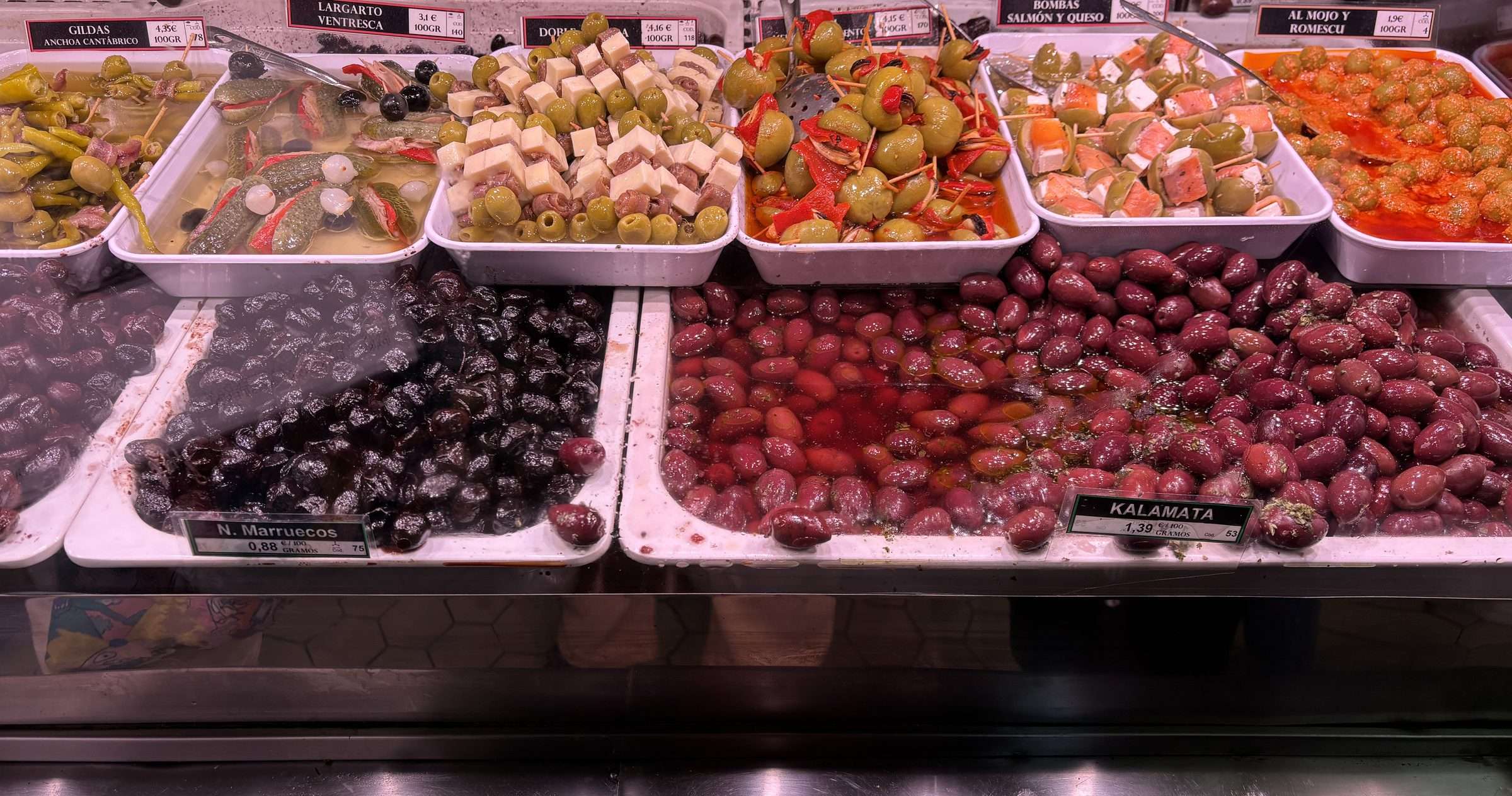 Bandejas de aceitunas y encurtidos con sus carteles de precio en un puesto de mercado