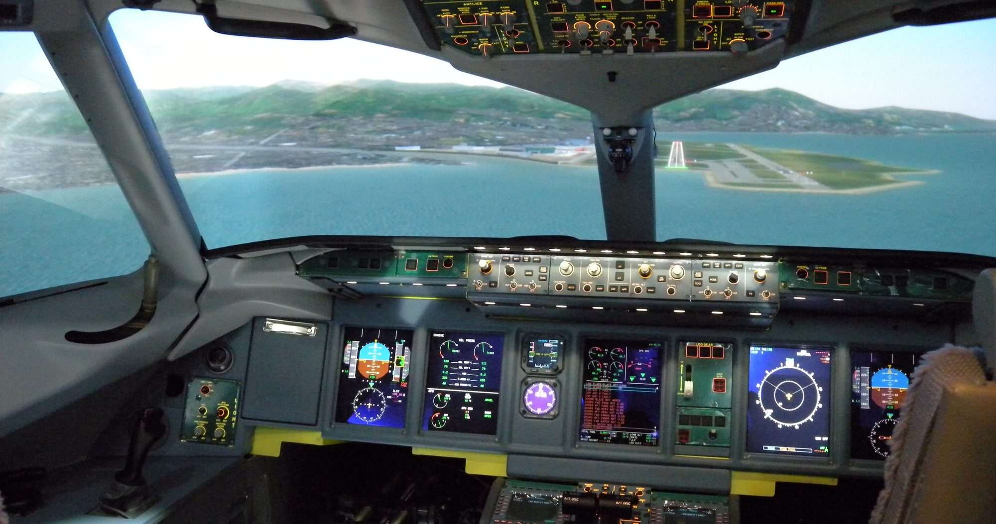 Cabina de un simulador de vuelo con las pantallas encendidas y una pista de aterrizaje junto al mar proyectada en las ventanas